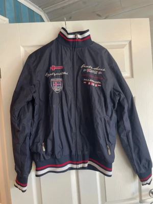 Marinblå vindjacka från Paul & Shark - Snygg marinblå vindjacka från Paul & Shark med broderade detaljer och patchar på bröstet. Jackan har ståkrage, dragkedja framtill och ribbade muddar med röd, vit och blå rand. Perfekt för dig som gillar sportig och nautisk stil.