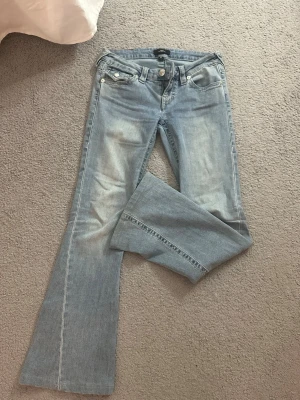 Trendiga ljusblå bootcut jeans från true religon low waist - Snygga ljusblå bootcut jeans från true religon med klassisk femficksmodell och silverfärgade detaljer. Jeansen har låg midja och är tillverkade i mjukt denimtyg som ger en avslappnad look. Jeansen är i modellen joey och bär storleken w25 (passar mig som är 162cm lång och ungefär 62-64cm midjemått). Jeansen ser sprillans nya ut och har inga defekter. Sitter väldigt bra i midjan och i längden! 