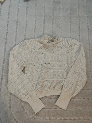 Vit spetsstickad tröja med puffärm - Supersöt vit stickad tröja med spetsmönster och puffärmar. Tröjan har hög hals med liten volang och breda ribbade muddar vid ärmslut och midja. Perfekt för dig som gillar romantisk och trendig stil.