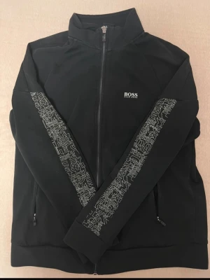 Hugo boss kofta  - Snygg svart ziptröja från BOSS med vit logga på bröstet och coolt grafiskt mönster längs ärmarna. Tröjan har hög krage, dragkedja framtill och två sidofickor med dragkedja. Tillverkad i mjukt material som känns bekvämt och stilrent.