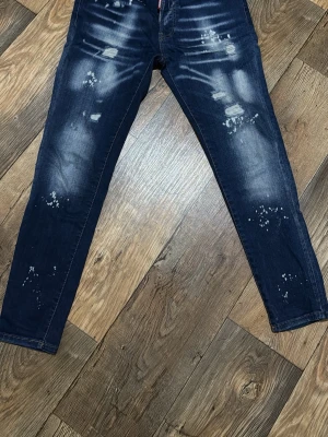 DSQUARED2 passar för dig som är 175cm - Säljer ett par mörkblå jeans med slitningar och vita färgstänk för en trendig, avslappnad look. Jeansen har smal passform och klassisk femficksdesign. Perfekta för dig som gillar streetstyle och vill sticka ut lite extra.