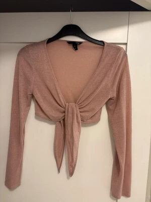 Glittrig beige croppad topp H&M - Säljer en glittrig brige bolero från H&M med lång ärm och djup v-ringning.Boleron har en snygg knytning framtill som ger en trendig look. Perfekt för dig som vill sticka ut lite extra med en feminin touch.