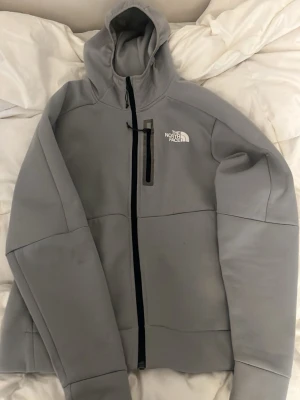 Grå kofta från The North Face - Snygg grå hoodie från The North Face med dragkedja framtill och en vertikal bröstficka med svart dragkedja. Hoodien har en stor huva och logga på både bröstet och ryggen. Tillverkad i ett mjukt och stretchigt material som känns skönt mot huden och är väldigt fin.