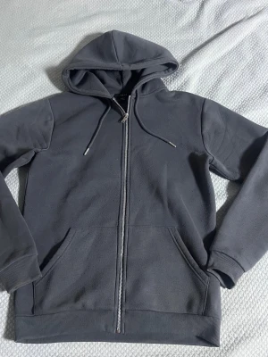 Mörkgrå zip hoodie med fickor - Snygg mörkgrå hoodie med dragkedja framtill och två stora fickor. Tröjan har huva med dragsko och ribbade muddar vid ärmar och nederkant. Perfekt för en avslappnad stil och enkel att matcha med andra plagg.