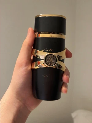 Versace parfym duo herr - Två parfymer från Versace: en svart och guld flaska med elegant design och Versace-logga i mitten, samt en blå glasflaska med texten 'Blue Jeans Man' i gult och detaljerad relief. Båda flaskorna har unika former och exklusiv känsla.