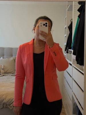 Koralrosa figursydd kavaj - Snygg korallrosa kavaj med figurnära passform och klassisk krage. Kavajen har två fickor med lock framtill och dekorativa knappar vid ärmsluten. Perfekt att styla över en enkel topp för en trendig look.