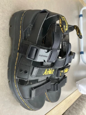 Svarta sandaler från Dr. Martens - Coola svarta sandaler från Dr. Martens med robust gummisula och justerbara remmar. Märkets klassiska gula söm och AirWair-logga i gult på remmen. Sandalerna har spännen och är tillverkade i slitstarkt syntetmaterial och läder. Mycket bra skick, får ingen rättvis bild på sandalerna.