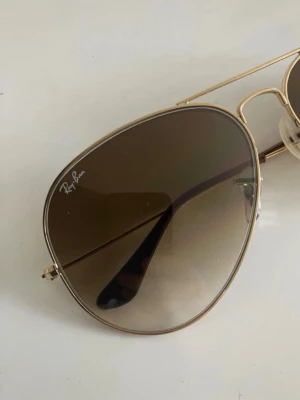 Ray-Ban aviator solglasögon guld - Snygga Ray-Ban aviator solglasögon med tunna guldfärgade metallbågar och bruna tonade glas. Klassisk pilotmodell som ger en cool vibe och passar till många stilar. Ikonisk Ray-Ban-logga på linsen och bekväma näskuddar.