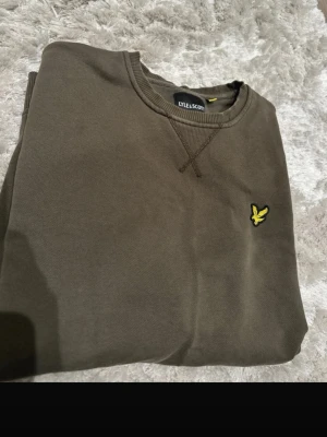 Olivgrön sweatshirt från Lyle & Scott - Snygg olivgrön sweatshirt från Lyle & Scott med rund halsringning och klassisk gul logga på bröstet. Tröjan har ribbade muddar vid ärmslut och nederkant samt en diskret detaljsöm vid halsen. Perfekt för en avslappnad och stilren look.
