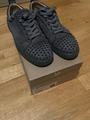 Svarta Louboutin sneakers med nitar - Säljer ett par svarta sneakers från Christian Louboutin i mocka med coola grå  nitar på tån. Skorna har klassisk låg profil, svarta skosnören och en stilren design som sticker ut. Perfekta för dig som vill ha något extra till din outfit. Sulerna är röda för en mer stilren look. Skick 7/10 lite smutsiga, storlek 46. Tveka inte att skriva vid minsta fundering