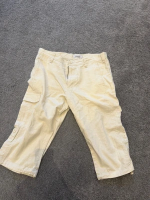 Beiga cargoshorts med fickor - Säljer ett par beiga cargoshorts med flera praktiska fickor på sidorna och knappar vid bensluten. Shortsens avslappnade passform gör dem perfekta för varma dagar. Klassisk dragkedja och bälteshällor i midjan.