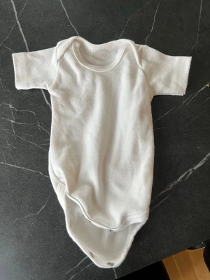 Vit kortärmad body - En enkel och stilren vit body med korta ärmar och rund hals. Praktisk knäppning med tryckknappar i grenen. Perfekt basplagg till garderoben. Materialet känns mjukt och bekvämt mot huden.