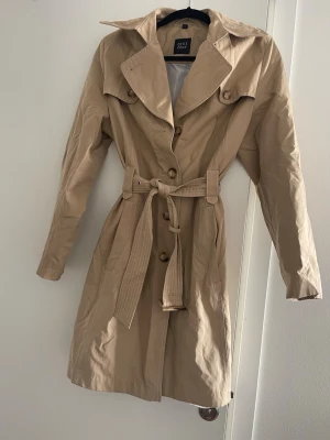 Beige trenchcoat - Stilren beige trenchcoat med bred krage och avtagbart midjebälte. Jackan har långa ärmar och knappar framtill. Knappt använd! 