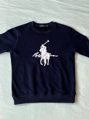 Mörkblå sweatshirt från Polo Ralph Lauren - Snygg mörkblå sweatshirt från Polo Ralph Lauren med stort vitt polospel-tryck på bröstet. Tröjan har rund halsringning, ribbade muddar vid ärmslut och nederkant samt en avslappnad passform. Perfekt för en stilren och sportig look.