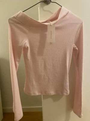 Ljusrosa ribbad offshoulder topp - Trendig ljusrosa ribbad topp med offshoulder-design och långa ärmar. Toppen har en smickrande, croppad passform och är tillverkad i mjuk bomullsmix som känns skön mot huden. Perfekt för dig som vill ha en enkel men snygg bas i garderoben.