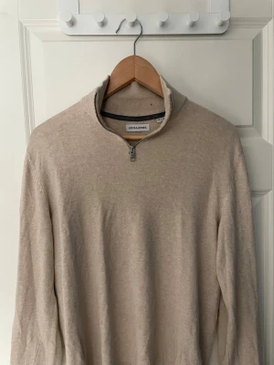 Beige halvzip-tröja från Jack & Jones - Beige stickad tröja från Jack & Jones med halv dragkedja och hög krage. Tröjan har en enkel och stilren design, perfekt för lager-på-lager. Materialet ser ut att vara mjukt och bekvämt, och modellen är långärmad. Storlek XS men passar S. 