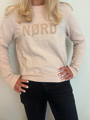 Beige sweatshirt med NØRD-tryck - Säljer en beige sweatshirt med trycket 'NØRD' framtill. Tröjan har rund halsringning, långa ärmar och ribbade muddar vid ärmslut och nederkant. Perfekt för en avslappnad och trendig look.