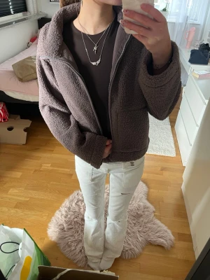 Brun teddyjacka med dragkedja - Mysig brun teddyjacka med dragkedja framtill och hög krage. Jackan har två stora fickor på sidorna och är gjord i fluffigt teddy-material som ger en avslappnad och trendig look. Perfekt för kyliga dagar och enkel att matcha med olika outfits.