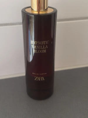 Zara Hypnotic Vanilla Bloom parfym - Zara Hypnotic Vanilla Bloom Eau de Parfum i en elegant mörkbrun cylinderformad flaska med gulddetaljer. Doften har en lyxig och modern känsla, perfekt för dig som gillar varma och söta vaniljtoner. Flaskan har stilren design med guldtext på framsidan.