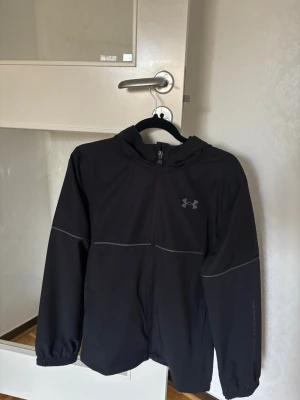 Svart vindjacka från Under Armour - Snygg svart vindjacka från Under Armour med huva och dragkedja framtill. Jackan har diskreta reflexdetaljer på armar och bröst samt elastiska muddar vid ärmsluten. Perfekt för blåsiga dagar och sportiga outfits. Köpte den för 800kr och vill sälja den för 700kr har inte använt den för att den är för stor