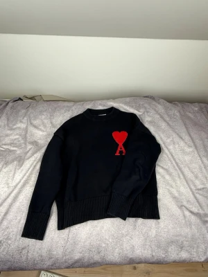 Svart stickad tröja från Ami - Säljer en svart stickad tröja från Ami Alexandre Mattiussi med röd hjärtlogga och bokstaven A på bröstet. Tröjan har ribbade muddar vid ärmslut och nederkant samt en klassisk rund halsringning. Perfekt för dig som gillar stilrena detaljer.