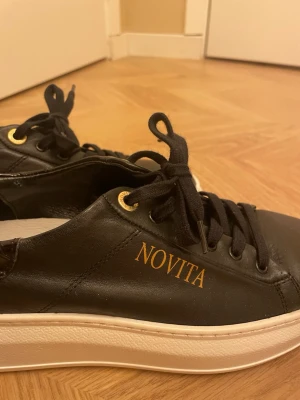 Svarta Novita sneakers i läder - Stilrena svarta sneakers från Novita i slätt läder med guldfärgade detaljer och logga på sidan. Skorna har svarta skosnören, vita tjocka sulor och guldfärgade öljetter som ger en lyxig känsla. Perfekta för dig som gillar klassisk men trendig stil.