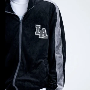 Tröja - En svart tröja med mörkgråa detaljer och grått tryck i tyg som står LA. Är något mjukare material som är riktigt skönt. Storlek L 