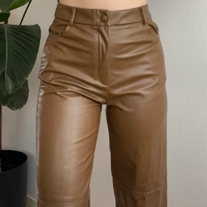 Byxor i brun läderimitation  - Byxor i brun läderimitation med knapp och dragkedja framtill och två fickor baktill. Rak modell som når ner till fötterna. Storlek L