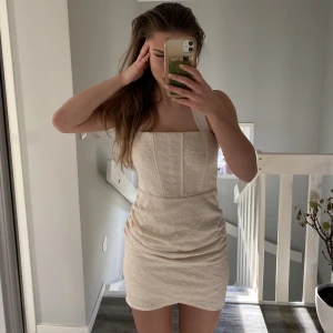 Krämvit jaquardklänning - Säljer denna jättefina jaquardvävda korsettklänningen från NaKd som har lite ”rush” framtill. Den har även knytband vid halsen så man kan justera den hur som helst. Har inte kommit till användning!🥰