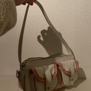 Handväska - Handväska, endast använd en gång. Vit med rosa detaljer, fickorna på framsidan går att öppna och använda. Bara att skriva vid frågor. Endast Swish och post.