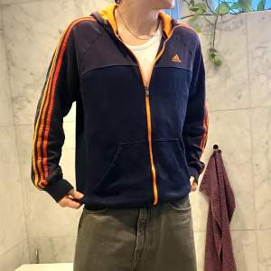 En mörkblå Adidas zip-up med orangea ränder, bra skick, knappt använd, nice passform