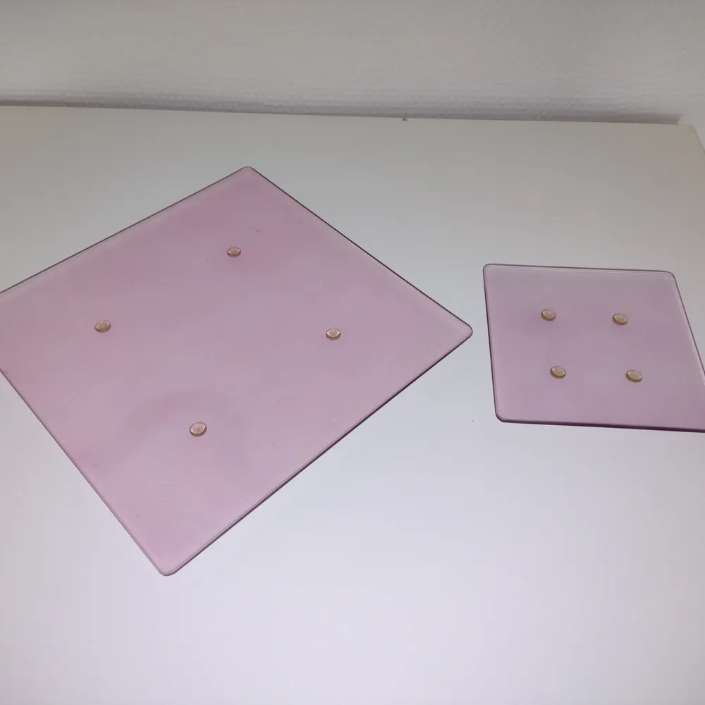 Två fyrkantiga rosa ljusfat/ljusbrickor/fat i glas. Det större är ca 20x20 och det mindre ca 11x11 cm stort. En del repor (märks inte av när de står på bordet men om man håller upp dem mot ljuset). Kan såklart användas till annat, som att lägga smycken på. Övrigt.