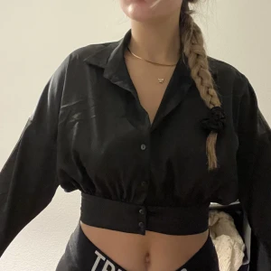 Glansig cropppad blus  - Denna svarta blus från bikbok har knappar och tightare band i midjan som får fram kroppen så snyggt 😍 den är i ett glansigt siden material och i strl xs 💖 helt oanvänd! 