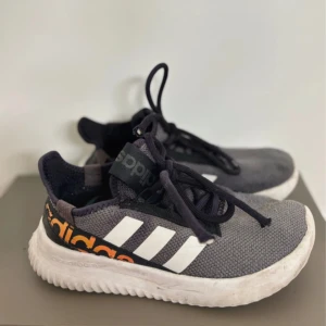 Gråa Adidas sneakers med Cloudfoam - Snygga grå sneakers från Adidas med klassiska vita ränder på sidan och orange Adidas-logga på hälen. Skorna har svart snörning och en bekväm vit Cloudfoam-sula. Ovandelen är i textil för en sportig look och skön känsla.