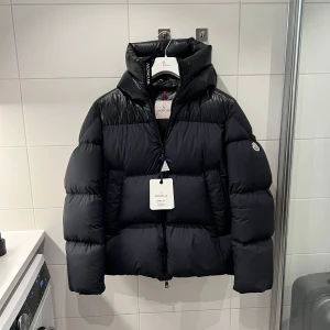 Moncler pufferjacka med huva - Oanvänd Moncler pufferjacka i en fluffig och mycket trendig modell. Perfekt för kalla dagar när du vill kombinera stil och funktion. Jackan har en generös vadderad huva, klassisk Moncler-logga på ärmen och är i nytt skick (endast testad) - Inga hål, fläckar eller slitage. Ettiketter är borttagna men finns kvar och har knutits fast igen. En exklusiv vinterjacka som håller dig både varm och stilsäker.