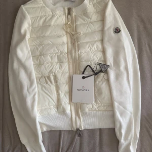 Vit quiltad dunjacka från Moncler - Snygg vit cardigan från Moncler med quiltad front och ribbstickade ärmar. Jackan har dragkedja framtill, två fickor och klassisk Moncler-logga på ärmen. Perfekt för dig som vill ha en clean och exklusiv look.