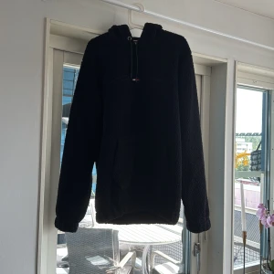 Svart fleece hoodie med huva - Mysig svart hoodie i fleece med huva och dragsko. Tröjan har en stor känguruficka framtill och långa ärmar. Perfekt när du vill vara både varm och chill. Passar dig som gillar en enkel och stilren look.