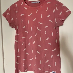 Rosa t-shirt med fjädermönster  - Söt rosa t-shirt från lager 157 i storlek XS, täckt av mönster med vita fjädrar. Använd fåtal gånger🩷