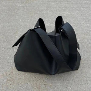 Svart väska - Stilren svart axelväska i slätt skinn med bred axelrem och minimalistisk design. Väskan har en mjuk, rymlig form och passar perfekt för dig som gillar enkel men snygg stil. Perfekt att slänga över axeln när du är på språng. Passar perfekt till skolan! Fint skick💖