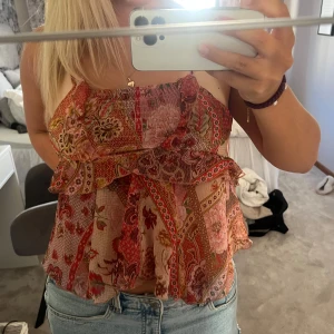 Blommig volangtop med tunna axelband - Supersöt blommig topp med volanger och tunna axelband. Toppen har ett färgglatt mönster i rött, rosa och beige med inslag av grönt och gult. Luftig och flowy passform, perfekt till jeans eller kjol. Materialet känns lätt och somrigt.