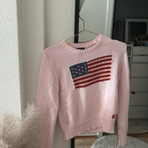Rosa stickad tröja Polo Ralph Lauren - Säljer en ljusrosa stickad tröja från Polo Ralph Lauren med amerikansk flagga framtill och RL-broderi vid ärmslutet. Tröjan har rund hals, ribbade muddar och lång ärm. Perfekt för dig som gillar preppy och klassisk stil.