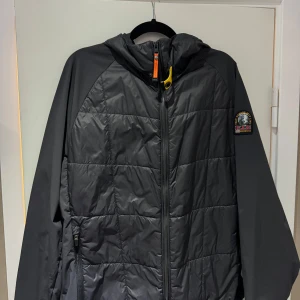 Svart pufferjacka från Parajumpers - Svart pufferjacka från Parajumpers med huva och coola detaljer som orange dragkedja och gul knapp vid halsen. Jackan har quiltad framsida, två fickor med dragkedja och klassisk PJS-logga på ärmen. Perfekt för dig som vill ha en stilren och funktionell jacka.