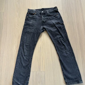 Levi's 501 svarta jeans W30 L30 - Klassiska svarta Levi's 501 jeans med raka ben och femficksmodell. Jeansen har en snygg tvättad svart färg, knappgylf och ikonisk läderpatch bak i midjan. Perfekta för dig som gillar tidlös stil och vill ha ett par jeans som funkar till allt. Använda med inga större defekter förutom hålet på vänster ben (se bild 5). 