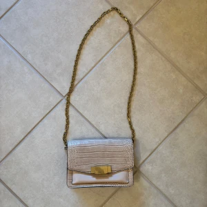 Beige axelväska från Marella - Snygg beige axelväska från Marella med krokopräglat mönster och guldfärgad kedja. Väskan har ett elegant spänne framtill och flera fack inuti, inklusive ett med dragkedja. Perfekt för dig som vill ha en stilren och trendig accessoar.