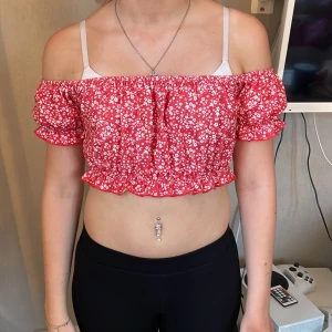 Röd blommig offshoulder croppad topp - Superfin röd croppad topp med vita småblommor och volangkant. Offshoulder-modell med puffiga korta ärmar och resår både upptill och nertill för en snygg passform. Perfekt för varma dagar och ger en söt och trendig vibe.