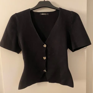 Svart topp från Gina Tricot - Svart v-ringad topp från Gina Tricot med korta ärmar och croppad passform. Toppen har snygga knappar framtill och är figursydd. Knappt använd, sitter dåligt på mig då de är en storlek för liten