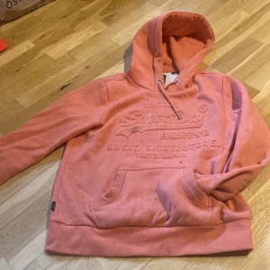 Superdry hoodie i korallorange - Säljer en snygg korallorange hoodie från Superdry med broderad logga framtill. Tröjan har huva med snörning, stor magficka och mjukt fleecefoder på insidan. Perfekt för dig som gillar streetstyle och vill ha något bekvämt och trendigt. Ska tvättas
