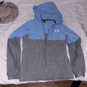 Blå och grå vindjacka från Under Armour - Snygg tvåfärgad vindjacka från Under Armour i ljusblått och grått. Jackan har huva, hel dragkedja framtill, lång ärm och logga på bröstet. Perfekt för dig som gillar sportig stil och vill ha något lätt och funktionellt till vardagen.