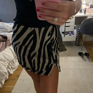 Zebramönstrad kortkjol med knappar - Säljer en snygg kortkjol med zebramönster i svart och beige. Kjolen har en rak passform och cool knappdetalj längs ena sidan. Perfekt för dig som vill sticka ut och addera lite edge till din outfit.