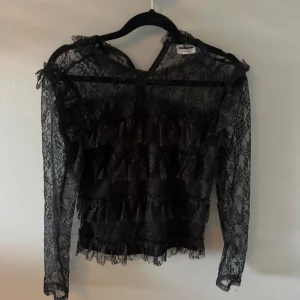 Svart spetsblus med volanger - Supersnygg svart blus från Boohoo i transparent spets med blommigt mönster. Långa ärmar och flera lager volanger framtill ger en cool och romantisk vibe. Perfekt för dig som vill sticka ut med detaljer och älskar spets. Har dock två små hål på ena armen. 